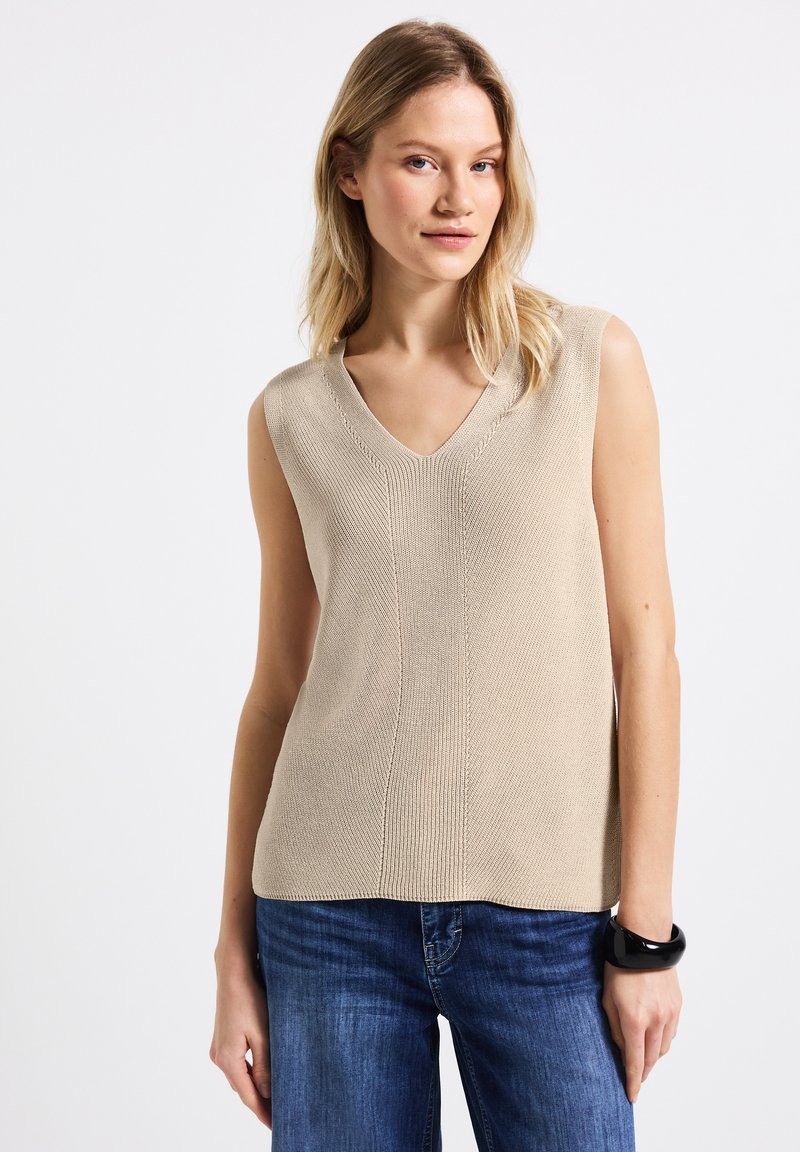 Top senza maniche beige in maglia con scollatura a V, caratterizzato da un design testurizzato e dettagli di cucitura verticale, abbinato a jeans in denim blu e un bracciale nero.