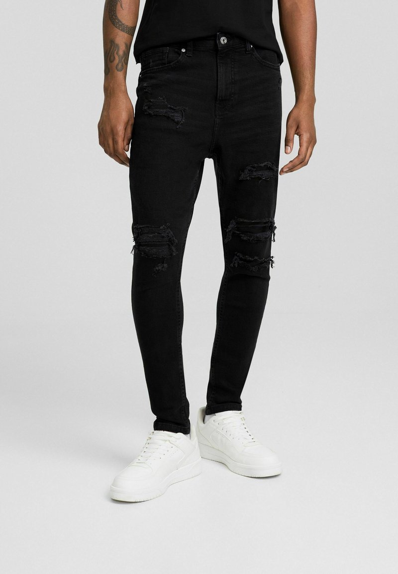 Vaqueros skinny de mezclilla negra con desgarros desgastados, que cuentan con un diseño clásico de cinco bolsillos y costuras tonales sutiles. Combinados con zapatillas blancas.
