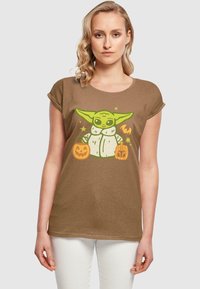 Brun kortärmad t-shirt med en grön karaktär som håller två Halloween-pumpor, med grafiska detaljer och en avslappnad passform.