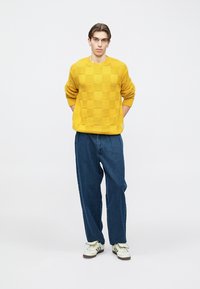 Žltý pletený sveter so šachovnicovým vzorom, doplnený voľnými modrými rifľovými nohavicami a bielymi teniskami.