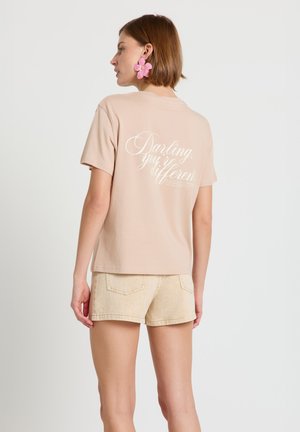 Donna che indossa una maglietta beige con testo bianco scritto sul retro e pantaloncini beige chiaro, rivolta verso sinistra, con orecchini a forma di fiore rosa.