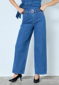 Pantaloni evazați din catifea bleu cu talie înaltă, prevăzuți cu o curea neagră cu accesorii metalice și o textură subtilă cu nervuri verticale.