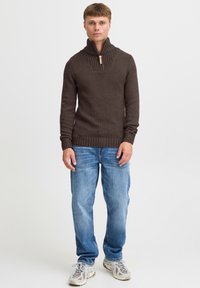 Brun strikket sweater med høj krave og vertikal ribbet tekstur, båret med lyseblå jeans og grå sneakers. Enkel design, afslappet pasform.