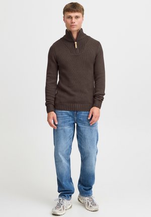 Brūns adīts džemperis ar augstu apkakli un vertikālu ribu tekstūru, valkāts kopā ar gaiši zilām džinsām un pelēkām kedām. Vienkāršs dizains, ikdienišķs piegriezums.