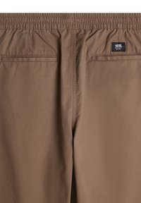 Bruine elastische broek met twee achterzakken, gestikte details en een geweven merklabel op de rechterzak. Zachte, gladde textuur.