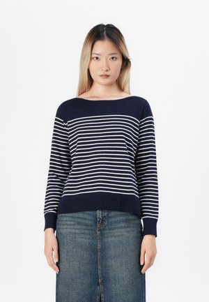 Maglione blu navy con strisce orizzontali bianche, scollo rotondo, maniche lunghe e orlo a costine. Abbinato a una gonna di jeans.