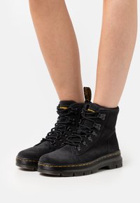 Botas de tobillo de ante negro con una suela de goma gruesa, que presentan ojales de metal, cordones negros y una etiqueta de marca amarilla en el tobillo.