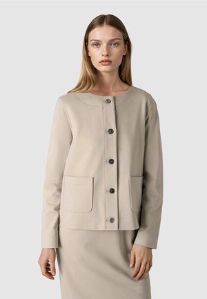 Frau mit glattem blondem Haar trägt eine beige Jacke mit großen Fronttaschen und passenden Rock, sie steht vor einem einfarbigen Hintergrund.