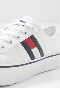 Sneaker bianca in tela con suola piatta, caratterizzata da strisce rosse e blu navy, occhielli metallici rotondi e lacci bianchi. Tessuto resistente.