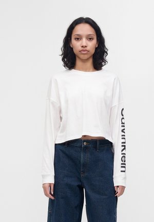 Junge Frau mit dunklen lockigen Haaren, die ein weißes, langärmeliges, bauchfreies Shirt mit dem Text "Calvin Klein" am Ärmel und blaue Jeans trägt.