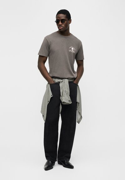 Άνδρας που φοράει γυαλιά ηλίου, γκρι μπλουζάκι με γραφικό σχέδιο, μαύρο παντελόνι και ασπρόμαυρο καρό πουκάμισο δεμένο γύρω από τη μέση, στέκεται με τα χέρια στις τσέπες.