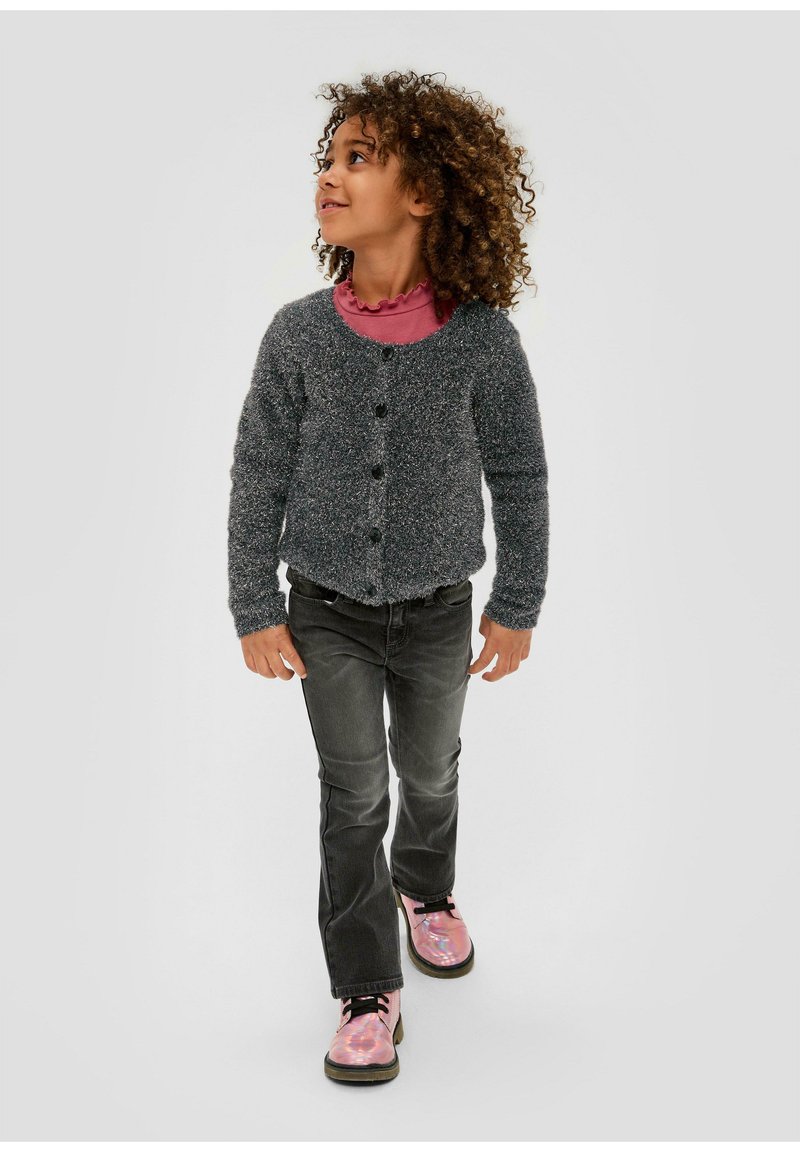 Cardigan gris duveteux avec des boutons noirs, porté sur une chemise rose à volants, associé à un jean foncé et des bottes roses brillantes avec des lacets noirs.