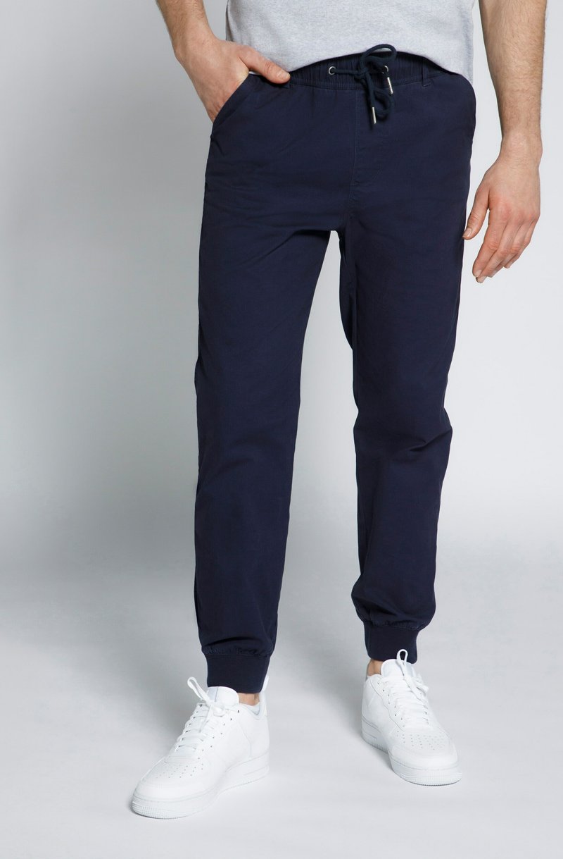 Donkerblauwe joggingsbroek met een trekkoord in de taille, zijzakken en elastische boorden, gecombineerd met witte sneakers op een lichte achtergrond.
