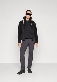 Crna fleece zip jakna s visokim ovratnikom, elastičnim vezicama, u kombinaciji s tamnosivim rastezljivim hlačama i crnim planinarskim cipelama.