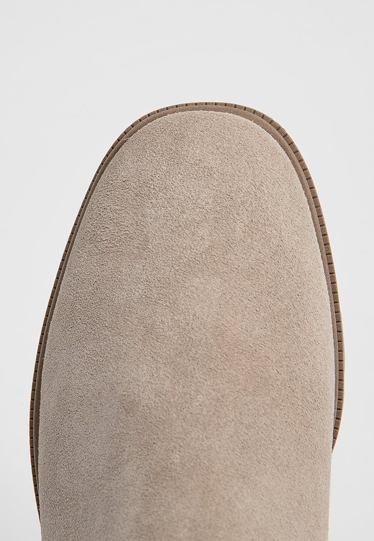 Beżowe zamszowe botki za kostkę z zaokrąglonym noskiem, subtelną teksturą i przeszyciami wzdłuż podeszwy i krawędzi.