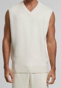 Pull sans manches en maille couleur crème, présentant un col en V, des bords côtelés et une finition texturée. Design minimaliste, tissu doux.