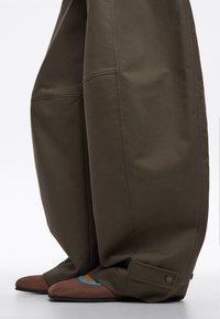 Pantalones oscuros de olivo anchos con puños abotonados, combinados con zapatos planos marrones que presentan un ribete azul brillante a lo largo de la abertura.