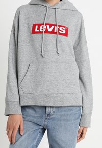 Felpa grigia con logo "LEVI'S" rosso e bianco. Presenta una tasca a marsupio frontale, cappuccio con cordino e polsini a coste. Tessuto morbido.