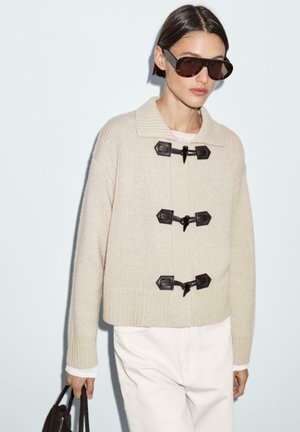 Beige knitted sweater with a wide collar, three dark toggle fasteners, ribbed hem, and long sleeves, paired with a white shirt and dark sunglasses.