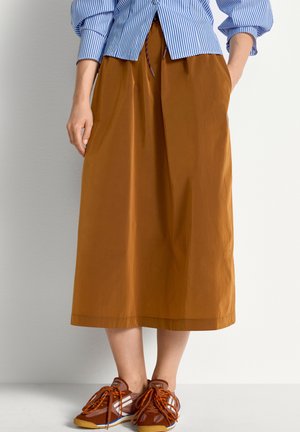 Femme portant une jupe midi couleur rouille avec des poches, associée à des baskets orange et marron et une chemise bleu à rayures rentrée.