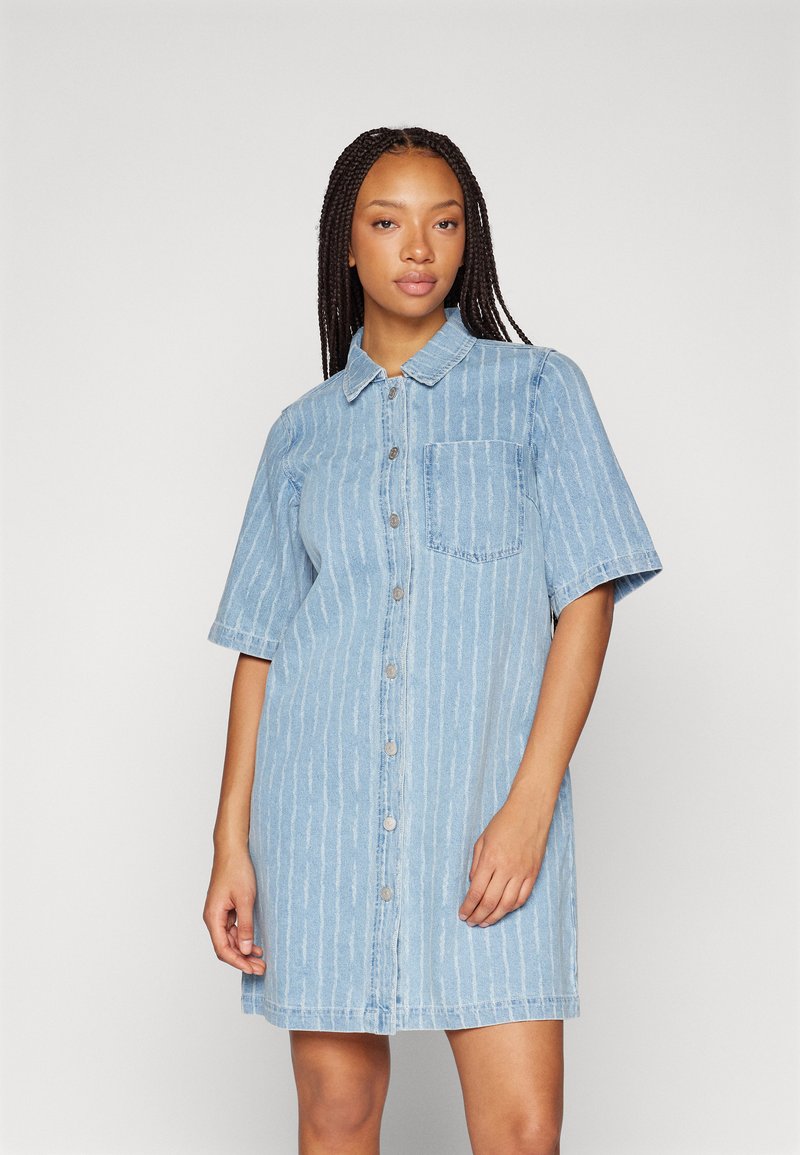 Lyseblå denimskjortekjole med korte ermer, knappestolpe foran og vertikalt stripemønster. Har en brystlomme og en avslappet passform.