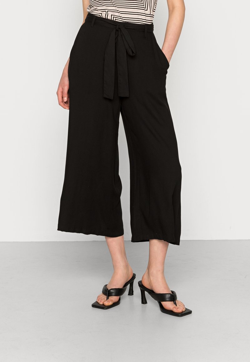 Pantaloni neri a gamba larga con cintura a cravatta, dotati di tasche laterali e tessuto morbido. Abbinati a sandali neri con tacco su uno sfondo neutro.