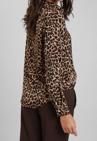 Blusa con stampa leopardata in tessuto setoso, dotata di colletto, polsini con bottoni e una vestibilità rilassata. Il motivo presenta macchie scure su uno sfondo beige.