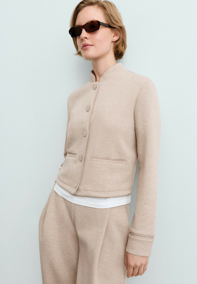 Femme portant une veste beige boutonnée et un pantalon assorti avec une chemise blanche en dessous, posant devant un mur gris clair, portant des lunettes de soleil foncées.