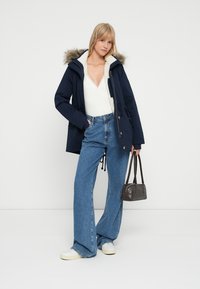 Parka blu con cappuccio foderato in pelliccia, bottoni frontali, grandi tasche, abbinato a un top in maglia bianco e jeans blu a vita alta, mentre tiene una piccola borsa.