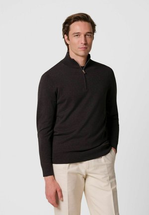 Uomo che indossa un maglione scuro a manica lunga con mezza cerniera e pantaloni beige chiaro, in piedi con una mano in tasca davanti a uno sfondo neutro.
