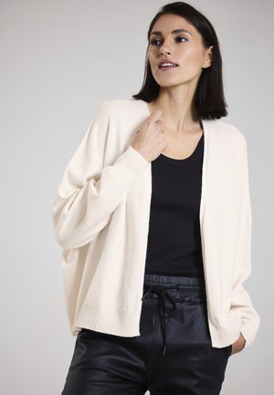 Helles beige Cardigan mit lockerer Passform, gerippten Bündchen und offener Front, getragen über einem schwarzen Tanktop und schwarzen Lederhosen. Glatte Textur.