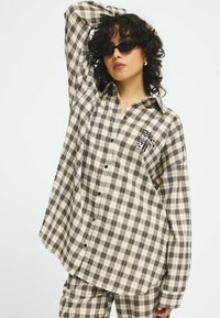 Oversized beige plaid shirt featuring black and brown checks, front button closure, and logo detail on the chest. Styled with dark sunglasses.