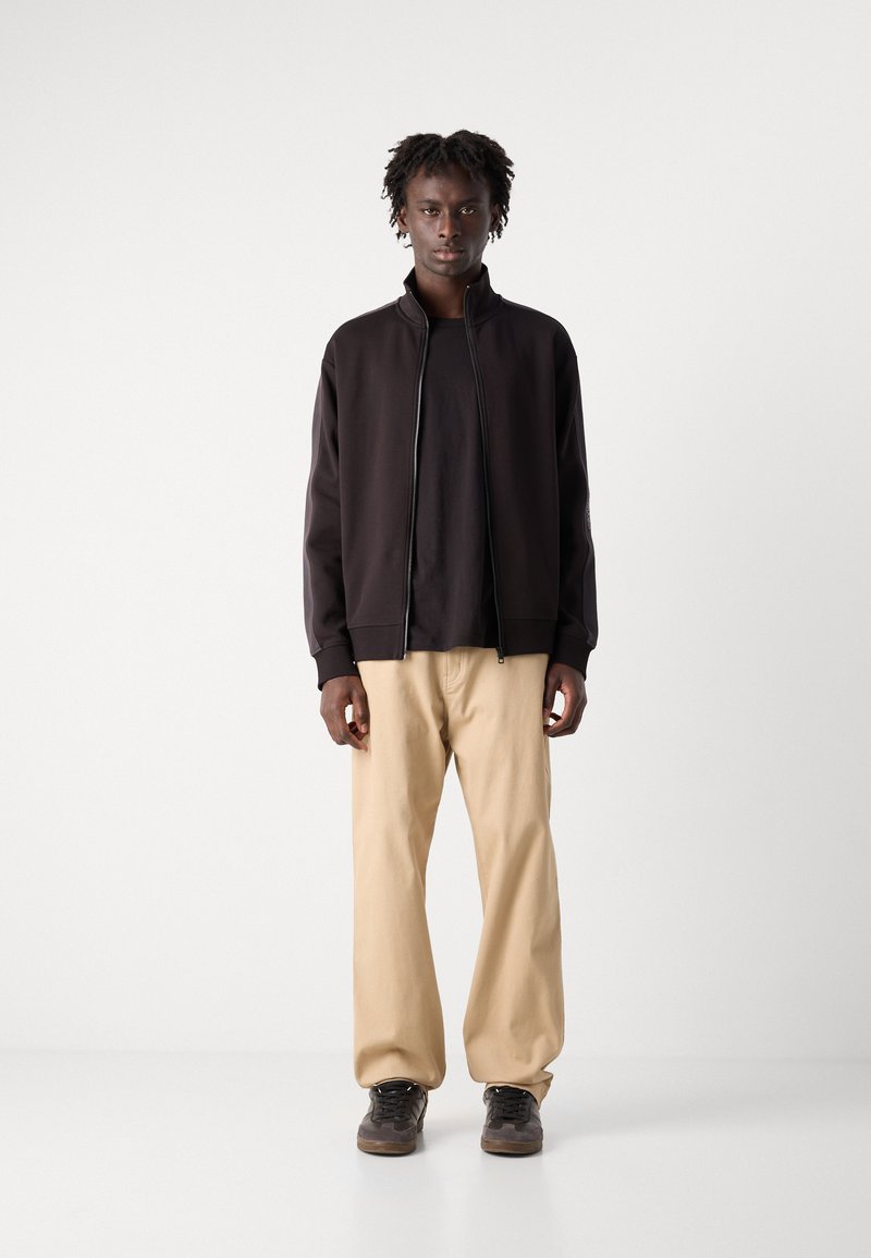 Felpa nera con zip sopra una maglietta nera, abbinata a pantaloni beige larghi e scarpe scure. Design semplice, senza motivi o loghi visibili.