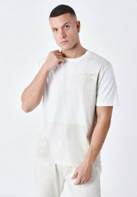 Camiseta de color claro con un diseño texturizado que presenta parches abstractos en gris claro. Cuenta con un sutil texto de la marca en el pecho. Mangas cortas.