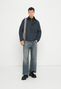 Giacca imbottita blu navy con chiusura a bottoni, indossata sopra un dolcevita verde oliva, abbinata a jeans larghi sbiaditi e scarpe nere chunky.