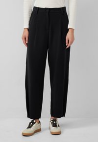 Pantalons noirs taille haute avec des plis, poches latérales et fermeture à bouton. Tissu lisse avec une coupe décontractée, associés à des baskets beiges.