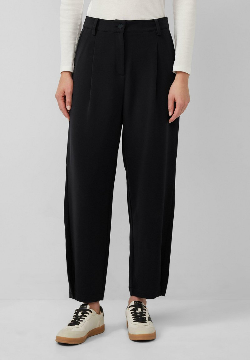 Pantalons noirs taille haute avec des plis, poches latérales et fermeture à bouton. Tissu lisse avec une coupe décontractée, associés à des baskets beiges.