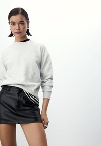 Grijze sweatshirt met lange mouwen, gecombineerd met een zwarte leren minirok met zijstrepen. Het model staat tegen een lichte achtergrond.