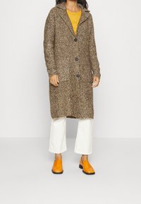 Brun, texturerad lång kofta med krage, prydd med stora knappar, bärs över en gul topp, tillsammans med vita byxor och orangea loafers.