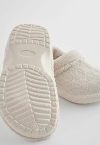 Des chaussons blancs et pelucheux avec une semelle en caoutchouc texturée. Design arrondi et upper doux ressemblant à de la laine. Une sangle au niveau de la cheville ajoute un détail pour le confort.