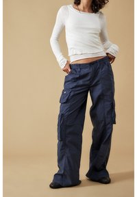 Person som bär en vit långärmad lagertröja och lösa marinblå cargobyxor med händerna i fickorna mot en beige bakgrund.