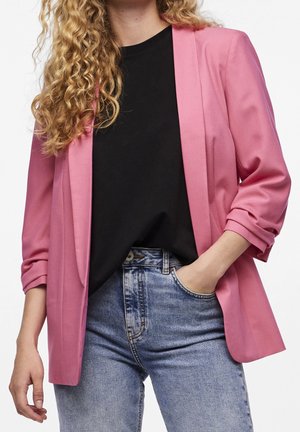 Kvinde med krøllet hår iført en pink blazer over en sort skjorte og lyseblå, højtaljede jeans, den ene hånd i lommen, foran en hvid baggrund.