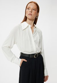 Weißes Langarmbluse mit spitzem Kragen und Knopfleiste, kombiniert mit hochgeschnittenen schwarzen Hosen und einem schwarzen Gürtel mit runder Schnalle.