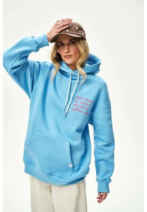 Kobieta w niebieskiej bluzie z kapturem z czerwonym napisem i brązową czapką z białymi motywami kwiatowymi, pozująca z jedną ręką na czapce.