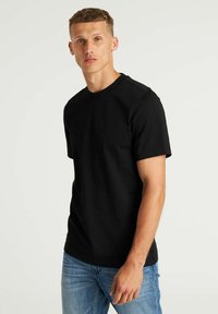 Crna pamučna majica s okruglim izrezom, kratkim rukavima i suptilnim kvadratnim logom na prsima. Glatka tekstura, prilagođen dizajn.