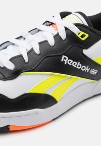 Černobílé tenisky se žlutými akcenty a texturovaným koženým svrškem. Mají bílé tkaničky, kulatou špičku a výrazné logo Reebok.