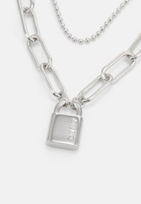 Sølvkjede med avlange ledd, prydet med en lås charm merket "DKNY" og akkompagnert av en mindre rund perlekjede.