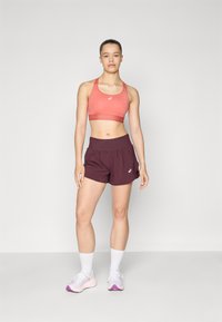 Sports bra in coral, matching shorts in dark burgundy, and white athletic socks. Model wearing light sneakers with purple accents.