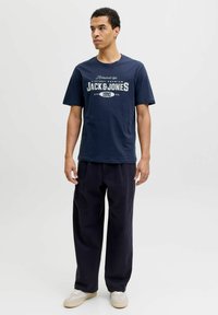 Camiseta de algodón azul marino con texto gráfico blanco "JACK & JONES 1990". Combinada con pantalones oscuros anchos y zapatillas ligeras sobre un fondo liso.