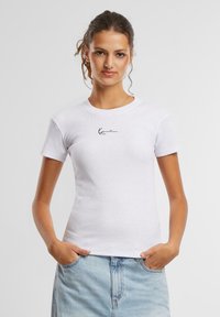 T-shirt blanche ajustée à manches courtes, texture côtelée, avec un logo noir signature sur la poitrine. Associé à un jean bleu clair.